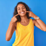 Woman with superb oral health and overall health in a yellow tanktop smiles and points to her teeth against a blue wall