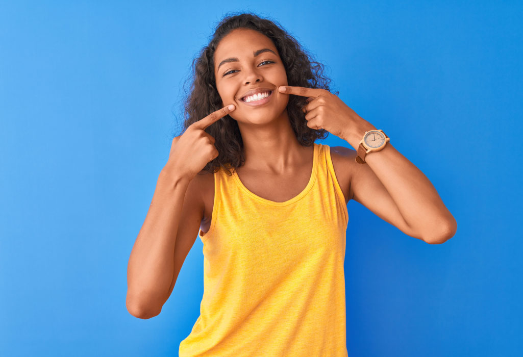 Woman with superb oral health and overall health in a yellow tanktop smiles and points to her teeth against a blue wall