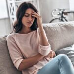 young woman sitting on the couch holding her head in pain from a migraine headache