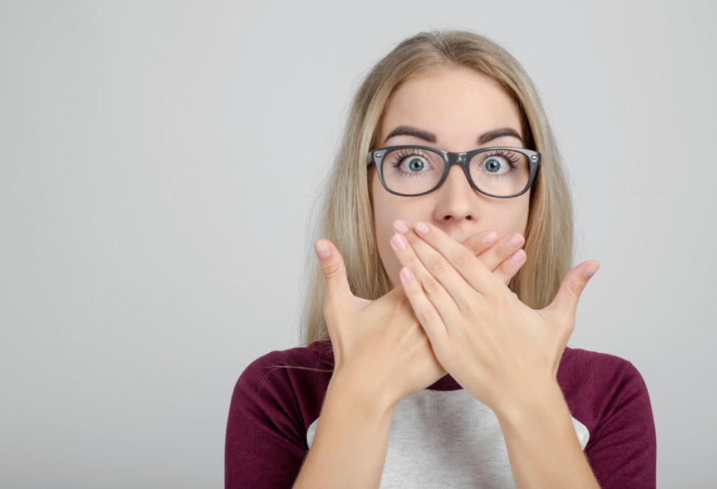 young woman wearing glasses covers her mouth with both hands to hide gum disease