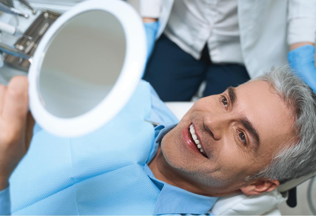 man in the dentist chair looks in a mirror at his restored gums and teeth.