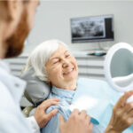 senior woman sitting in the dentist chair holds a mirror and looks at her implant supported dentures as the dentist looks on