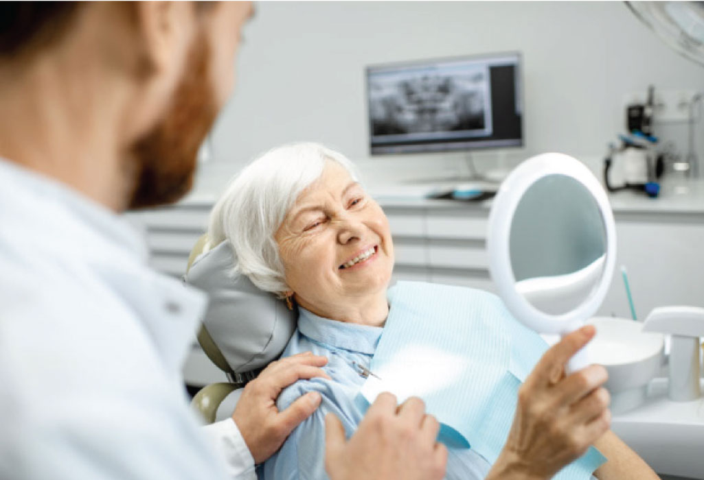 senior woman sitting in the dentist chair holds a mirror and looks at her implant supported dentures as the dentist looks on