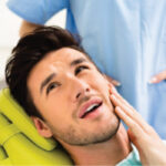 young man in the dentist chair holds his cheek with root canal pain